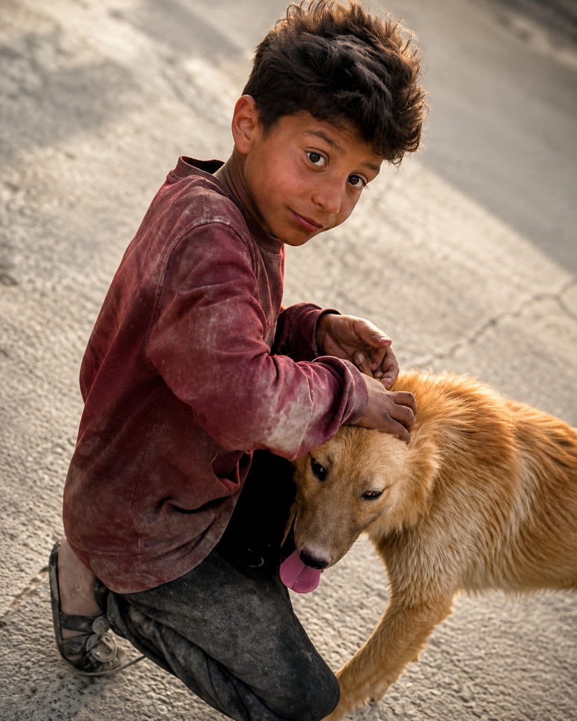 A boy crouching in the street with his arm around a dog